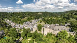 Panoramablick auf Monschau mit der Burg im Vordergrund, umgeben von grüner Landschaft und bewaldeten Hügeln unter blauem Himmel mit Wolken., © Eifel Tourismus GmbH, Dominik Ketz