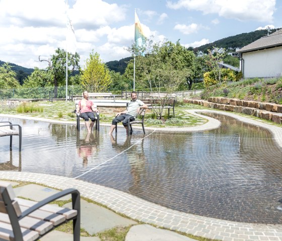 Zwei Personen sitzen entspannt auf Stühlen in einem flachen Wassergarten. Im Hintergrund sind Bäume, Büsche und ein Haus zu sehen., © Eifel Tourismus GmbH, AR-shapefruit AG