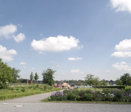 Grüne Parklandschaft im Seepark Zülpich mit Blumenbeeten, einem Weg und blauem Himmel mit weißen Wolken., © Nordeifel Tourismus GmbH
