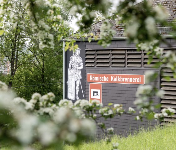 Gebäude mit Schild 'Römische Kalkbrennerei' und römischer Soldatengrafik, umgeben von Bäumen und Blüten., © Eifel Tourismus GmbH AR-shapefruit AG