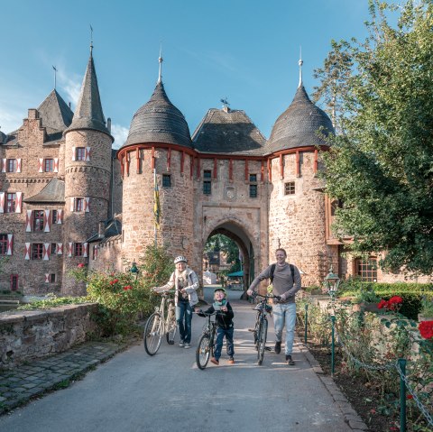 Eine Familie mit Fahrrädern steht vor der historischen Burg Satzvey. Die Burg hat markante Türme und rote Fensterläden. Es ist ein sonniger Tag., © Paul Meixner
