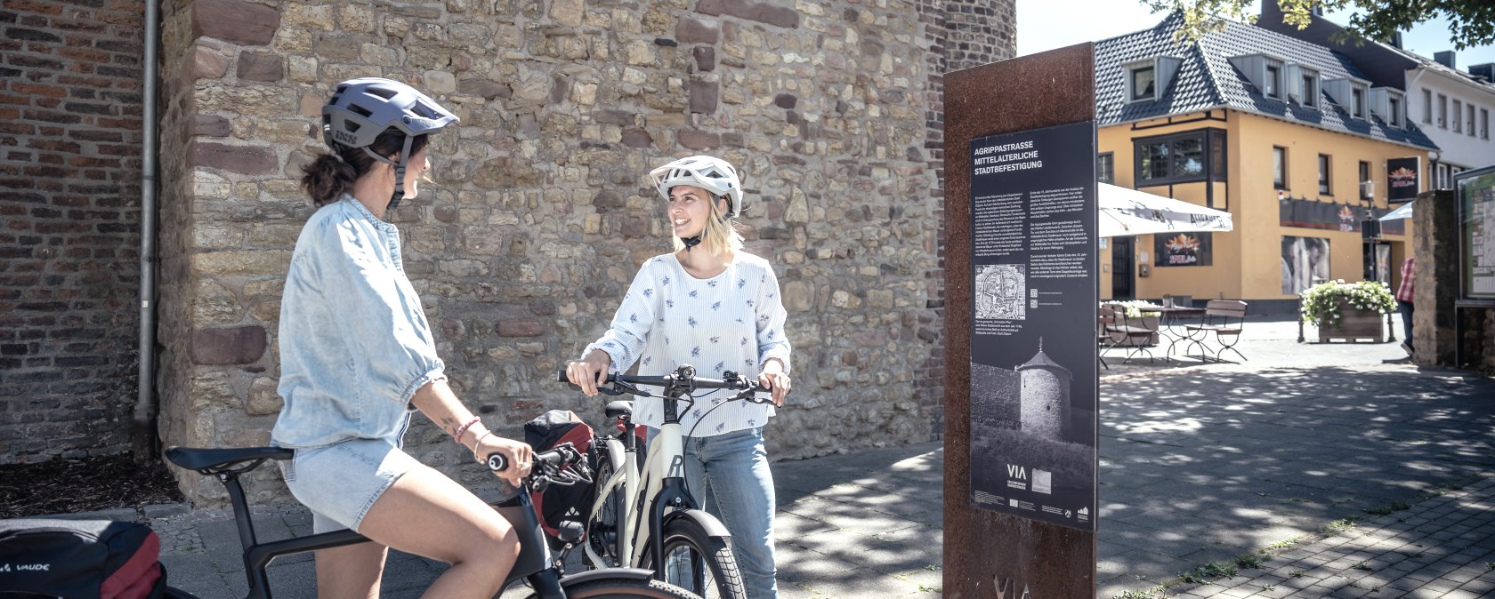 Zwei Fahrradfahrer mit Helm stehen in der Zülpicher Innenstadt vor dem Münstertor. Neben den Fahrradfahrern steht ein Infoschild., © Eifel Tourismus GmbH, Dennis Stratmann