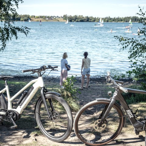 Zwei Fahrräder stehen am Ufer des Wassersportsees Zülpich. Zwei Personen blicken auf das Wasser, wo Segelboote und Surfer zu sehen sind., © Eifel Tourismus GmbH, Dennis Stratmann