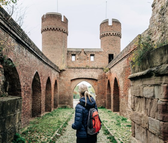 Eine Person mit rotem Rucksack steht vor einem alten Backsteinbogen mit Türmen im Park am Wallgraben in Zülpich., © PaulMeixner