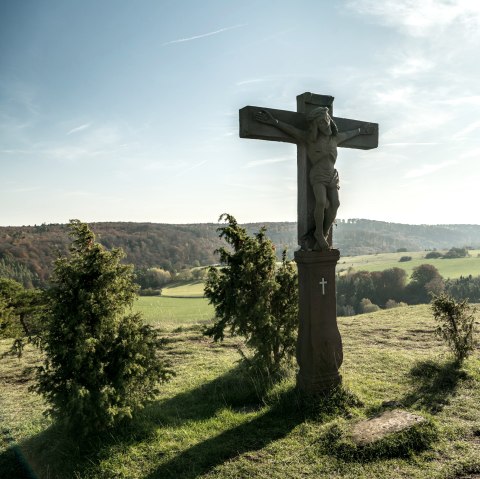 Ein Kreuz auf einem Hügel mit einer Bank daneben, umgeben von grüner Landschaft und bewaldeten Hügeln unter blauem Himmel., © Eifel Tourismus, D. Ketz
