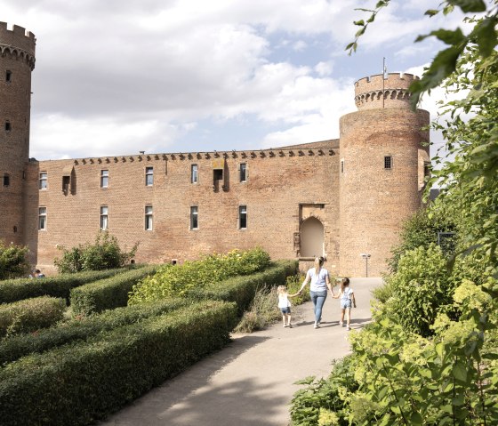 Historische Burg mit zwei T&uuml;rmen, umgeben von gepflegtem Garten. Eine Frau mit zwei Kindern spaziert auf einem Weg. Bew&ouml;lkter Himmel im Hintergrund., &copy; Eifel Tourismus GmbH, Tobias Vollmer