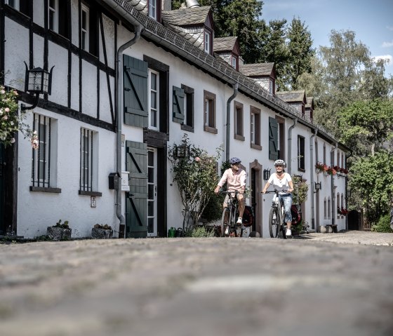 Zwei Radfahrer in einer malerischen Stra&szlig;e mit Fachwerkh&auml;usern in Kronenburg. Die H&auml;user sind mit Blumen geschm&uuml;ckt, und die Stra&szlig;e ist gepflastert., &copy; Eifel Tourismus GmbH, Dennis Stratmann