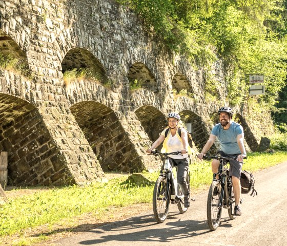 Zwei Radfahrer fahren auf einem sonnigen Weg entlang einer alten Steinmauer mit B&ouml;gen, umgeben von gr&uuml;ner Vegetation., &copy; Eifel Tourismus GmbH, Dominik Ketz