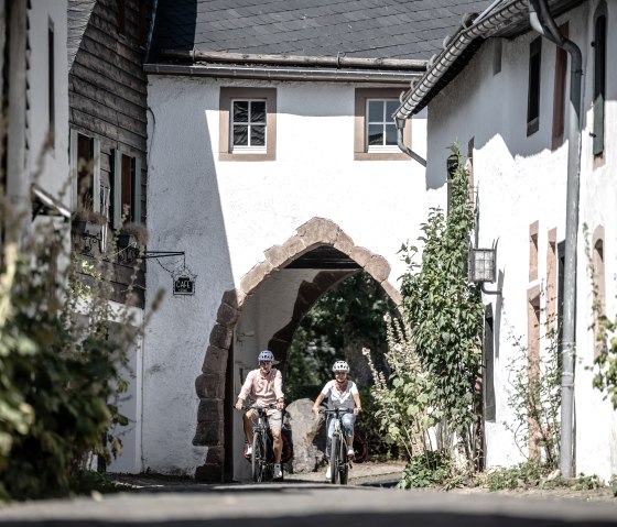 Two cyclists wearing helmets ride through a group of houses in the historic castle village of Kronenburg., &copy; Eifel Tourismus GmbH, Dennis Stratmann