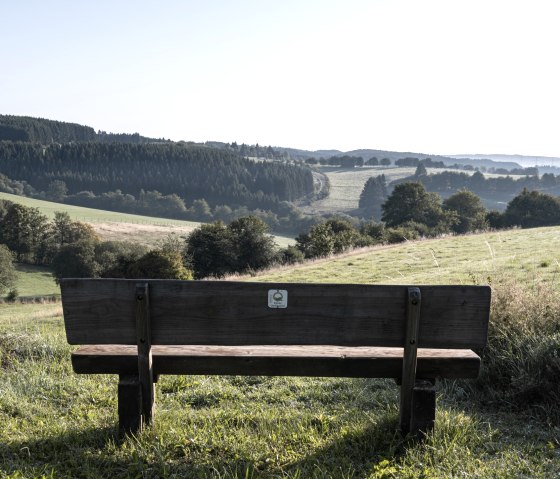 Eine Holzbank steht auf einer Wiese mit Blick auf die h&uuml;gelige Landschaft der Eifel, umgeben von W&auml;ldern und Feldern unter klarem Himmel., &copy; Nordeifel Tourismus GmbH