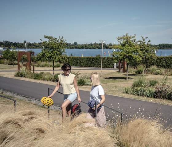 Zwei Personen spazieren im Seepark Z&uuml;lpich entlang eines Weges. Im Hintergrund ist der See und gr&uuml;ne Landschaft zu sehen., &copy; Eifel Tourismus GmbH, Dennis Stratmann