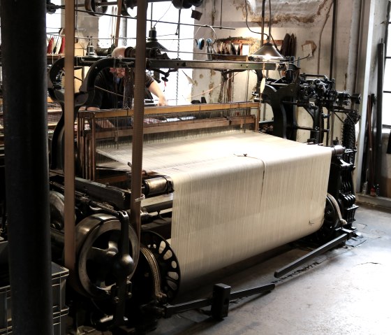 Ein alter Webstuhl in der LVR Industriemuseum-Tuchfabrik M&uuml;ller. Eine Person arbeitet daran, umgeben von industrieller Ausstattung., &copy; Nordeifel Tourismus GmbH