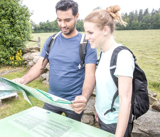 Two people with rucksacks look at a hiking map in front of a green meadow and trees., © Eifel Tourismus GmbH, AR-shapefruit AG