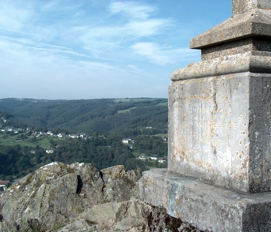 Steinernes Denkmal im Vordergrund, dahinter eine gr&uuml;ne H&uuml;gellandschaft mit einem Dorf unter blauem Himmel., &copy; Rursee-Touristik GmbH