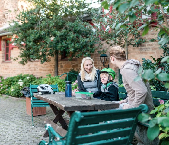 Family sitting at a wooden table in the courtyard of Satzvey Castle. Bicycles are parked next to them, with brick walls and plants in the background., &copy; Paul Meixner