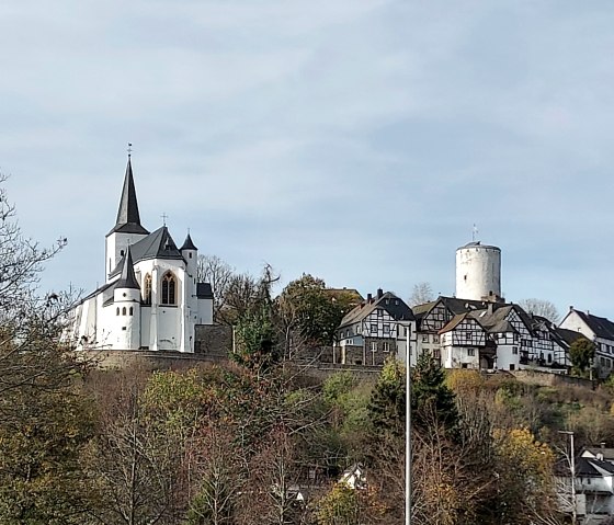 Blick auf die Burg Reifferscheid mit einem weißen Turm und umliegenden Fachwerkhäusern, umgeben von herbstlicher Vegetation., © Sweco GmbH