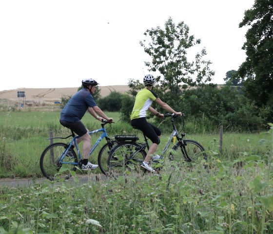 Zwei Radfahrer mit Helmen fahren auf einem Weg durch eine grüne, ländliche Landschaft mit Bäumen und Feldern., © Nordeifel Tourismus GmbH