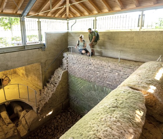 Zwei Personen stehen in einer überdachten römischen Brunnenstube bei Kallmuth und betrachten die antiken Strukturen., © Eifel Tourismus GmbH, Dominik Ketz