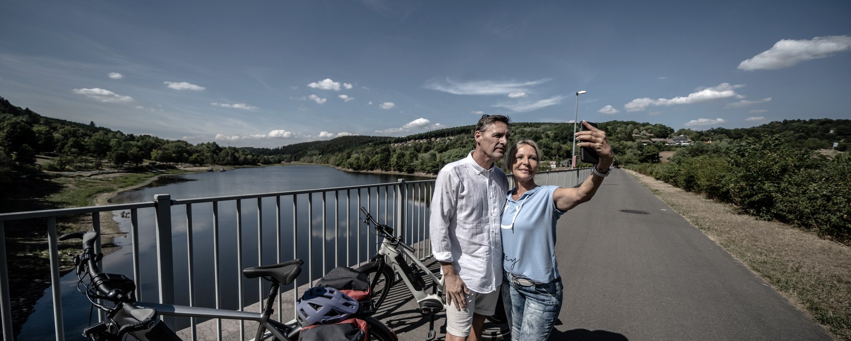 Ein Paar macht ein Selfie auf einer Brücke am Kronenburger See. Im Hintergrund sind Fahrräder und der See zu sehen., © Eifel Tourismus GmbH, Dennis Stratmann