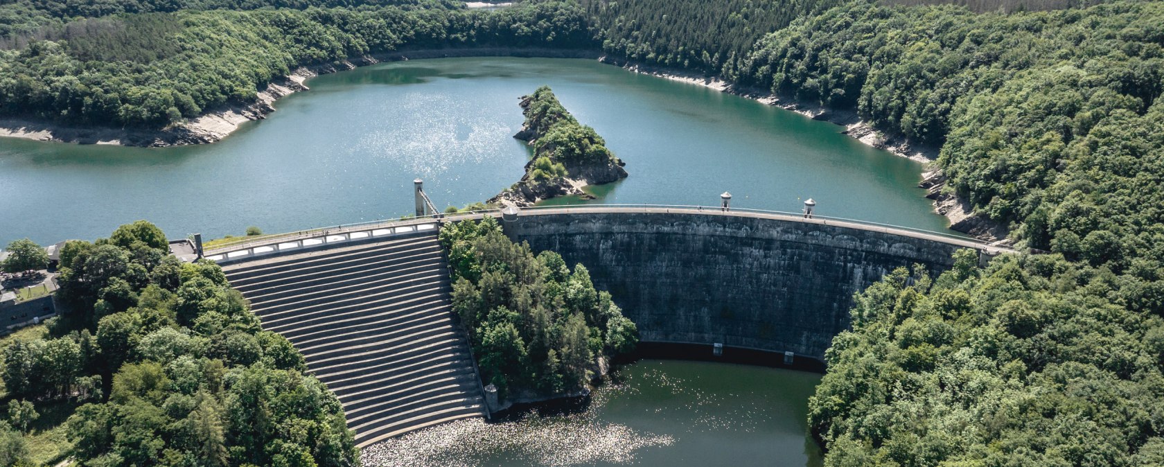Aerial view of the Urft dam with the Urft dam wall, surrounded by densely wooded hills and blue water under a clear sky., &copy; Eifel Tourismus GmbH, Dennis Stratmann - finanziert durch REACT-EU