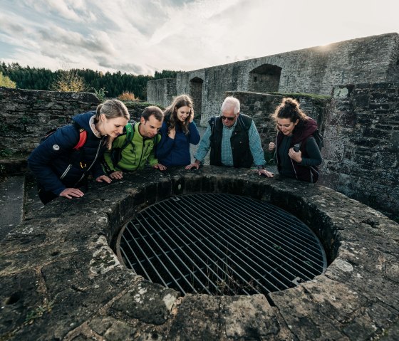 Eine Gruppe von fünf Personen blickt in einen mit einem Gitter abgedeckten Brunnen auf der Burg Reifferscheid. Im Hintergrund sind alte Mauern zu sehen., © Paul Meixner
