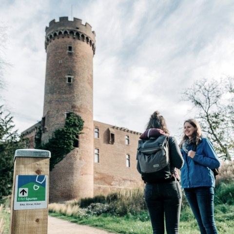 Zwei Personen vor einem historischen Turm in Z&uuml;lpich. Ein Wegweiser der Eifelspur steht im Vordergrund. Der Himmel ist bew&ouml;lkt., &copy; Paul Meixner