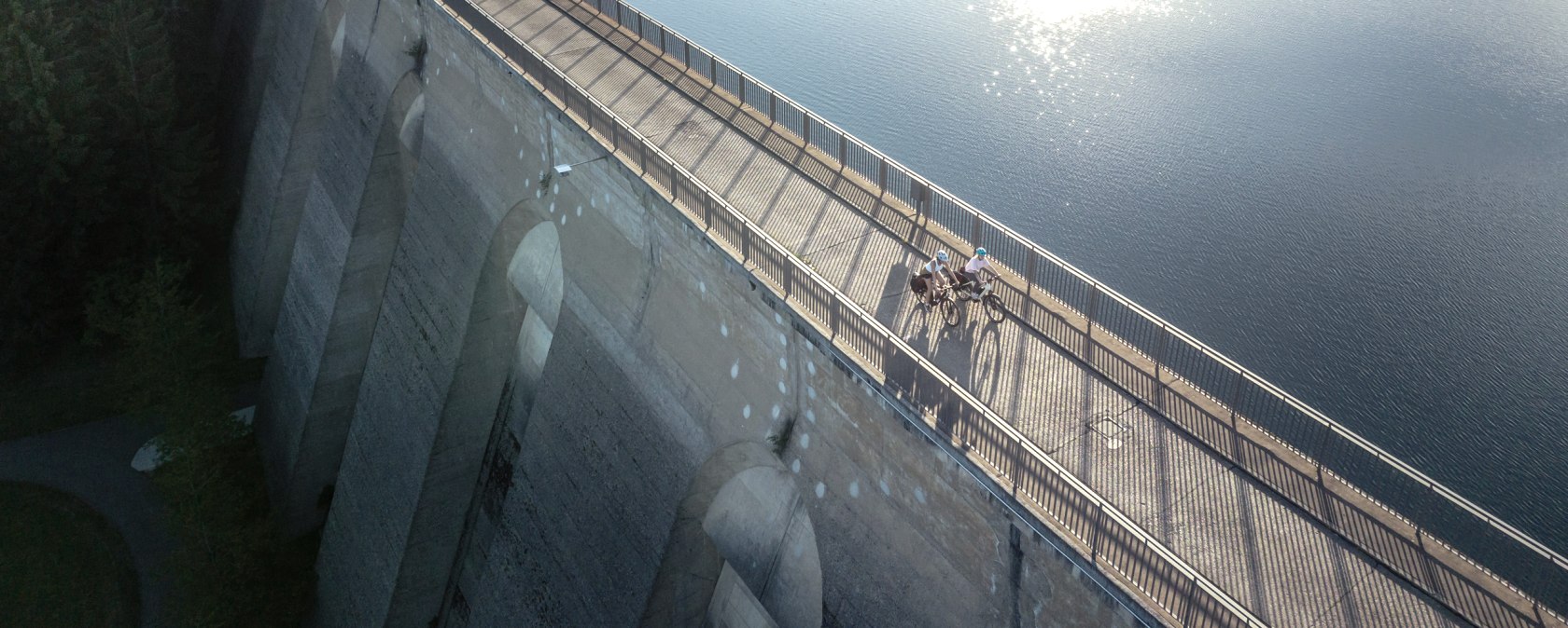 Luftaufnahme der Oleftalsperre in Hellenthal. Zwei Radfahrer fahren über die Staumauer, während die Sonne auf das Wasser scheint., © Eifel Tourismus GmbH, Dennis Stratmann