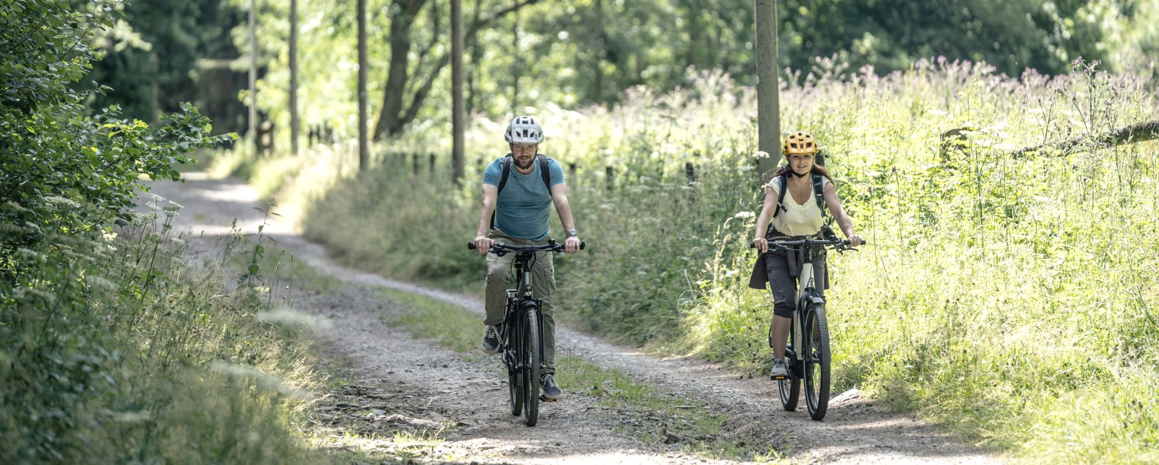 Zwei Radfahrer mit Helmen fahren auf einem sonnigen Waldweg im Prethbachtal, umgeben von grüner Vegetation., © Eifel Tourismus GmbH, Dennis Stratmann