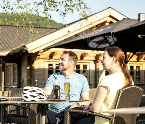 Zwei Personen sitzen entspannt an einem Tisch im Freien, genie&szlig;en Getr&auml;nke. Ein Fahrradhelm liegt auf dem Tisch. Im Hintergrund ein Holzgeb&auml;ude., &copy; Eifel Tourismus GmbH, Dominik Ketz