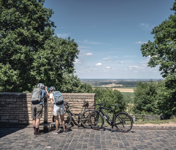 Zwei Radfahrer mit Helmen stehen an einem Aussichtspunkt in der Eifel. Sie blicken durch ein Fernglas auf die weite Landschaft. Fahrräder lehnen an der Mauer., © Eifel Tourismus GmbH, Dennis Stratmann