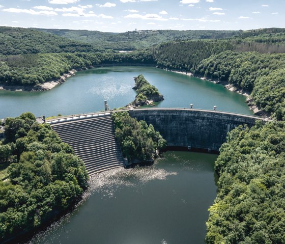 Luftaufnahme der Urfttalsperre mit der Urftstaumauer, umgeben von dicht bewaldeten H&uuml;geln und blauem Wasser unter einem klaren Himmel., &copy; Eifel Tourismus GmbH, Dennis Stratmann - finanziert durch REACT-EU
