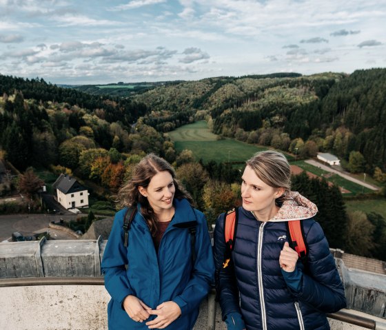 Zwei Frauen in blauen Jacken stehen auf einer Aussichtsplattform mit Blick auf eine grüne, bewaldete Landschaft und ein Dorf im Tal., © Paul Meixner