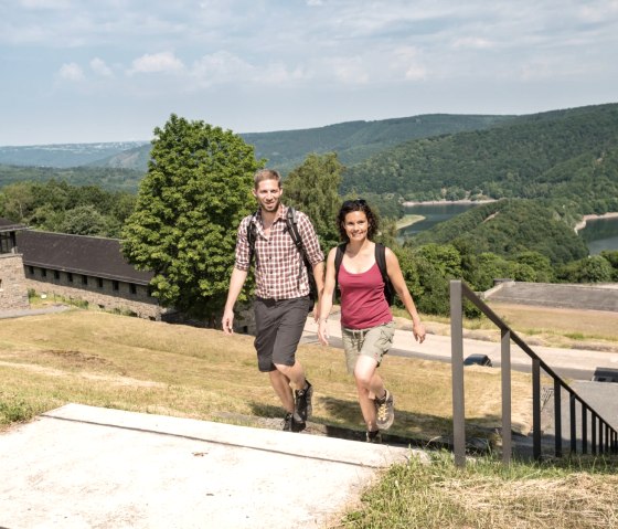 Zwei Wanderer steigen eine Treppe hinauf, im Hintergrund Vogelsang IP und gr&uuml;ne H&uuml;gel. Der Himmel ist leicht bew&ouml;lkt, die Stimmung sommerlich., &copy; Eifel Tourismus GmbH/D. Ketz
