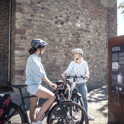 Zwei Fahrradfahrer mit Helm stehen in der Z&uuml;lpicher Innenstadt vor dem M&uuml;nstertor. Neben den Fahrradfahrern steht ein Infoschild., &copy; Eifel Tourismus GmbH, Dennis Stratmann