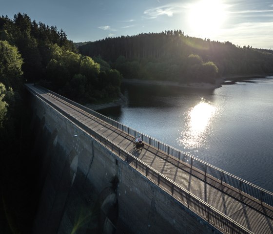 In the foreground, two cyclists are riding on a dam in sunny weather. In the background, the Oleftalsperre, on which the sun is reflected, is surrounded by a forest., &copy; Eifel Tourismus GmbH, Dennis Stratmann