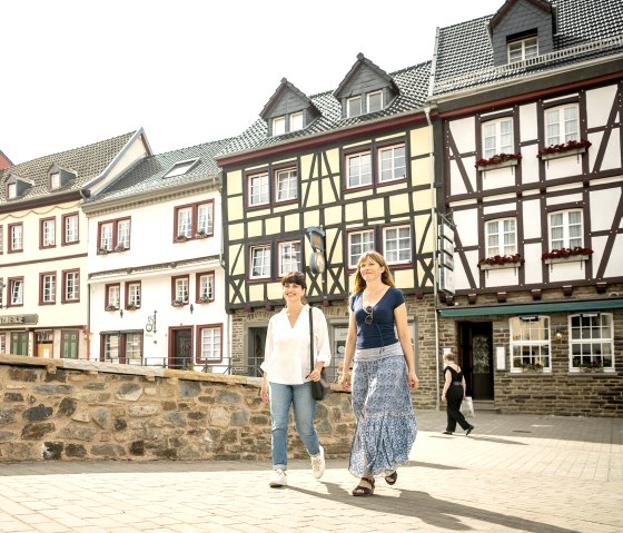 Zwei Frauen gehen lächelnd vor einer Reihe von Fachwerkhäusern in Bad Münstereifel entlang. Die Sonne scheint, und die Straße ist gepflastert., © Eifel Tourismus GmbH, Dominik Ketz