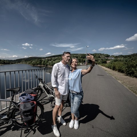 A couple takes a selfie on a bridge on Lake Kronenburg. Bicycles and the lake can be seen in the background., &copy; Eifel Tourismus GmbH, Dennis Stratmann