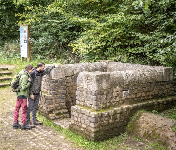 Zwei Personen betrachten einen alten Brunnen aus Stein in einem Waldgebiet. Im Hintergrund sind B&auml;ume und ein Informationsschild zu sehen., &copy; Eifel Tourismus GmbH, Dominik Ketz