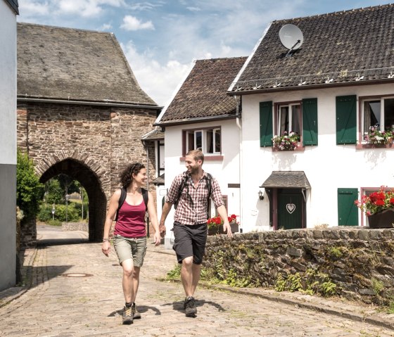 Ein Paar wandert durch den historischen Burgbering Reifferscheid, vorbei an Fachwerkhäusern und einem Torbogen., © Eifel Tourismus GmbH/D. Ketz