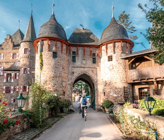 Satzvey Castle with two cyclists riding through the gate. The castle is surrounded by blooming roses and a clear blue sky., &copy; Paul Meixner