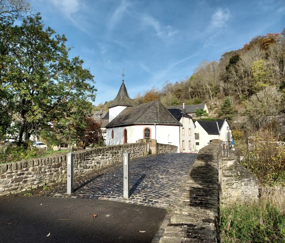 The St. Brigida Chapel stands on a cobbled path, surrounded by trees and hills under a blue sky., &copy; Sweco GmbH