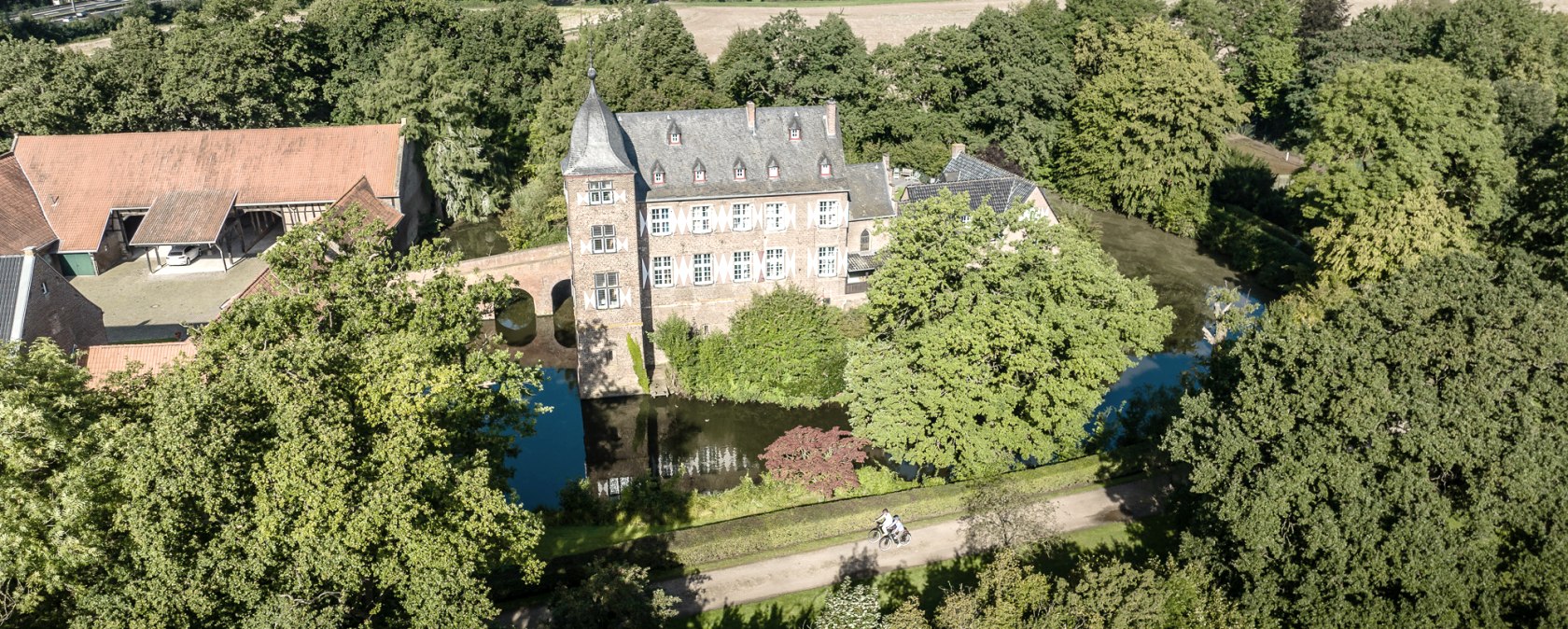 Luftaufnahme einer Burg mit Wassergraben, umgeben von B&auml;umen und Feldern. Ein Weg f&uuml;hrt an der Burg vorbei, auf dem zwei Radfahrer zu sehen sind., &copy; Eifel Tourismus GmbH, Dennis Stratmann