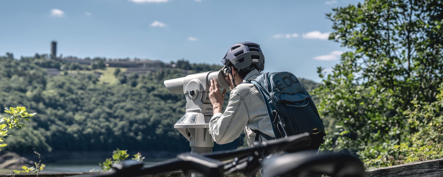 Person mit Helm und Rucksack schaut durch ein Fernglas. Im Hintergrund ist Vogelsang IP auf einem bewaldeten H&uuml;gel zu sehen., &copy; Eifel Tourismus GmbH, Dennis Stratmann