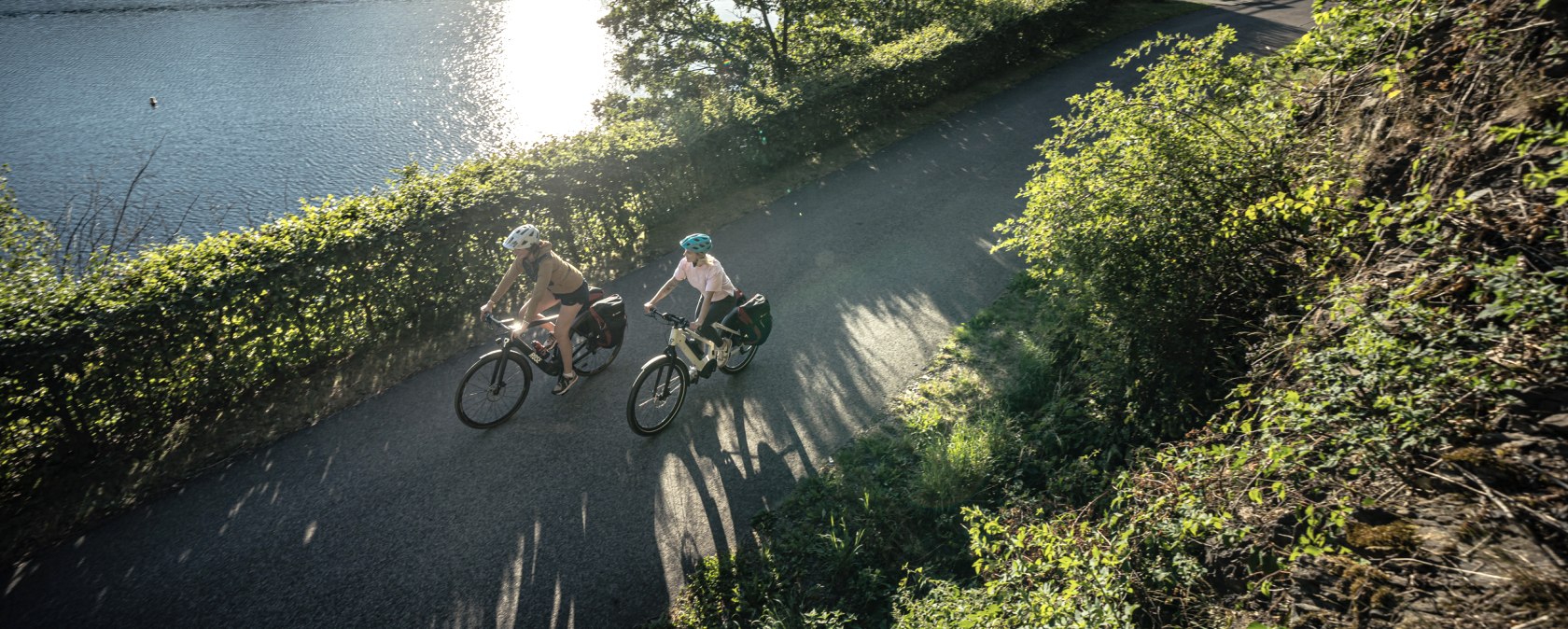 Zwei Radfahrer fahren bei Sonnenschein entlang eines Sees, umgeben von grüner Natur. Die Sonne spiegelt sich im Wasser., © Eifel Tourismus GmbH, Dennis Stratmann