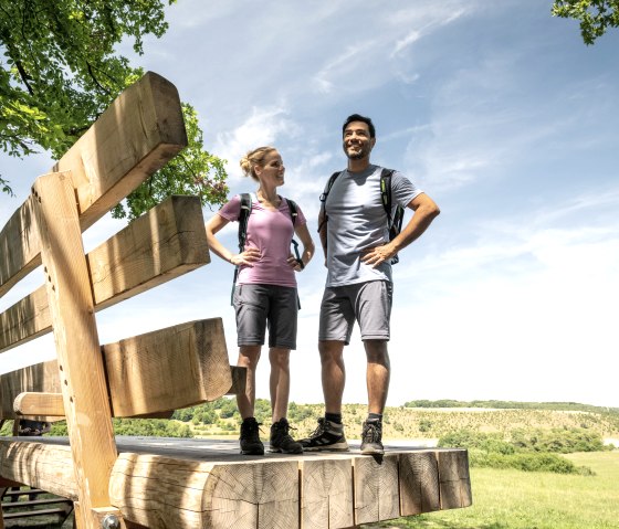 Zwei Personen stehen auf einer &uuml;bergro&szlig;en Holzbank in einer gr&uuml;nen Landschaft unter blauem Himmel. Sie tragen Wanderkleidung und Rucks&auml;cke., &copy; Eifel Tourismus GmbH, Dominik Ketz
