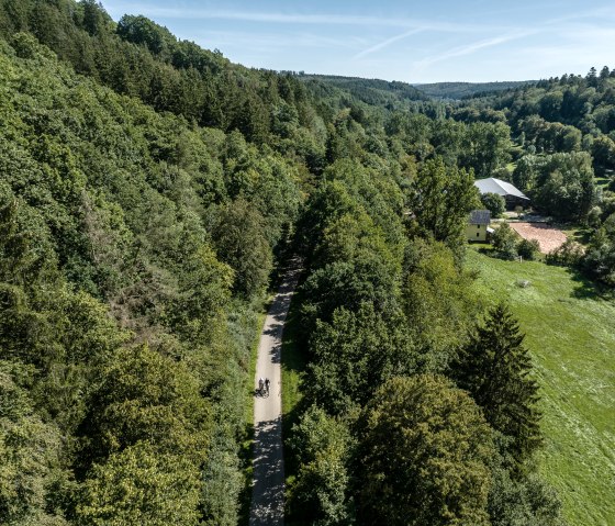 Luftaufnahme eines gr&uuml;nen Waldes mit einer Stra&szlig;e, auf der zwei Radfahrer fahren. Rechts ein Haus und eine Wiese, im Hintergrund H&uuml;gel., &copy; Eifel Tourismus GmbH, Dennis Stratmann