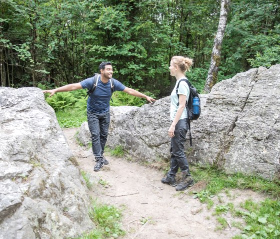 Zwei Wanderer stehen zwischen großen Felsen im Wald und unterhalten sich. Sie tragen Rucksäcke und Wanderkleidung., © Eifel-Tourismus GmbH, shapefruit AG A. Röser