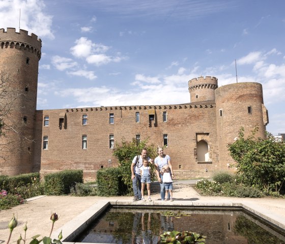 Eine Familie steht vor der Landesburg Zülpich. Im Vordergrund ist ein Teich, im Hintergrund die Burg mit Türmen und Zinnen., © Eifel Tourismus GmbH, Tobias Vollmer