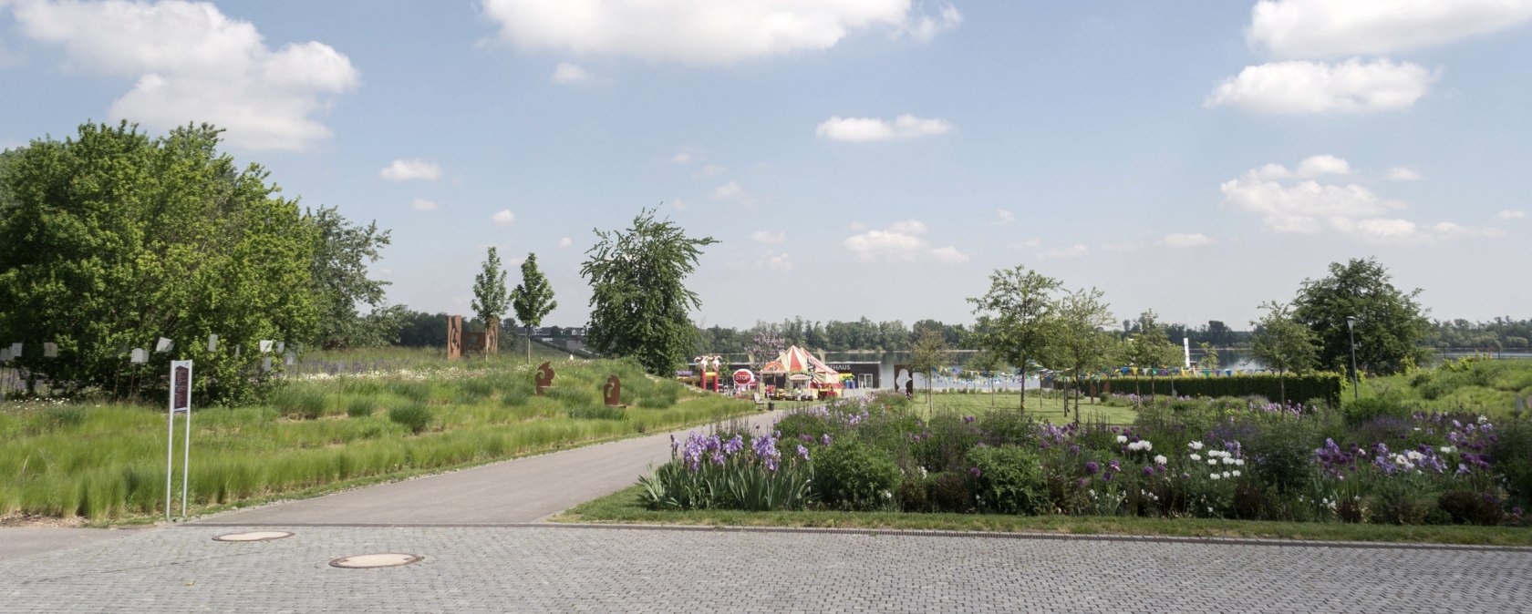 Grüne Parklandschaft im Seepark Zülpich mit Blumenbeeten, einem Weg und blauem Himmel mit weißen Wolken., © Nordeifel Tourismus GmbH