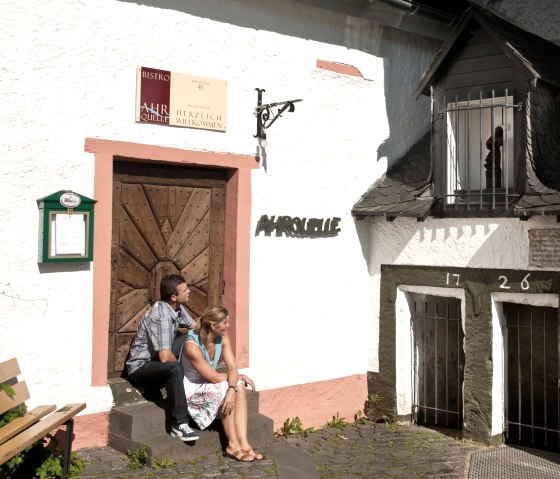 Zwei Personen sitzen vor der Ahrquelle in Blankenheim. Eine Holzt&uuml;r und ein kleines Fenster sind zu sehen. Die Jahreszahl 1726 ist an der Wand., &copy; Eifel Tourismus GmbH, D. Ketz